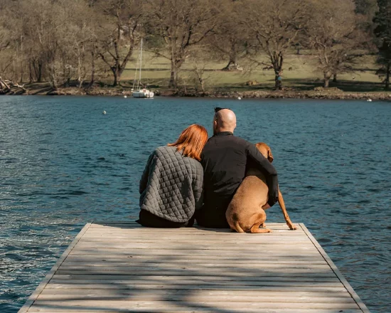 A young couple sit on the end of the Lakeside jetty overlooking Lake Windermere, with their dog.